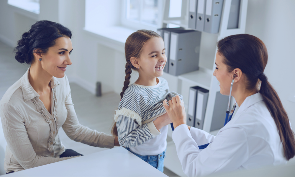 Image of a young girl with brown hair high-fiving her female doctor during a medical appointment, with her mother watching and smiling. The setting is a modern doctor's office, illustrating a supportive and positive healthcare environment for children. The doctor is dressed in a white lab coat, suggesting a professional and caring interaction.