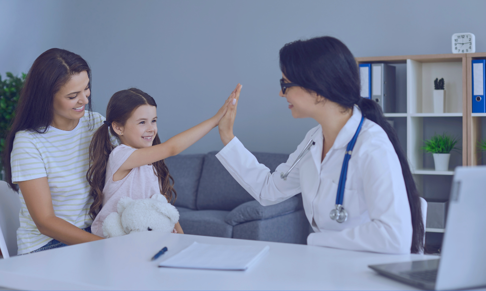 Image of a caring nurse in a dark blue uniform gently tending to a young boy lying in a hospital bed. The nurse is smiling at the boy, who is holding a teddy bear, creating a comforting atmosphere in the clinical setting. The room is well-lit with natural light streaming through large windows, enhancing the serene environment.