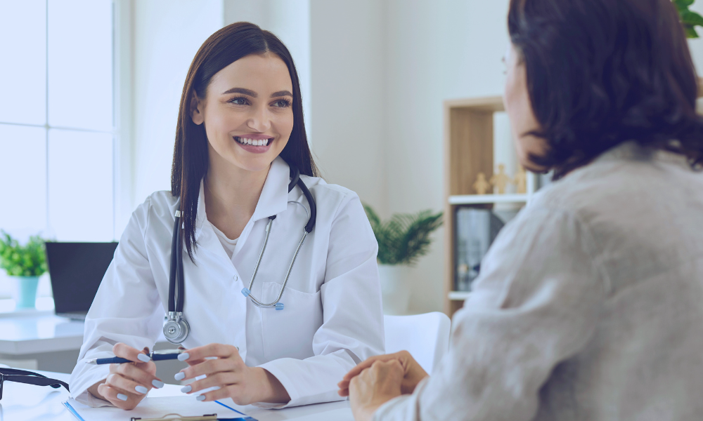 Image of a young boy in a hospital bed hugging his nurse in a comforting embrace. The nurse, wearing a light blue uniform, smiles warmly at the child, expressing care and support. They are in a brightly lit hospital room with large windows in the background, adding a serene and hopeful atmosphere to the scene.