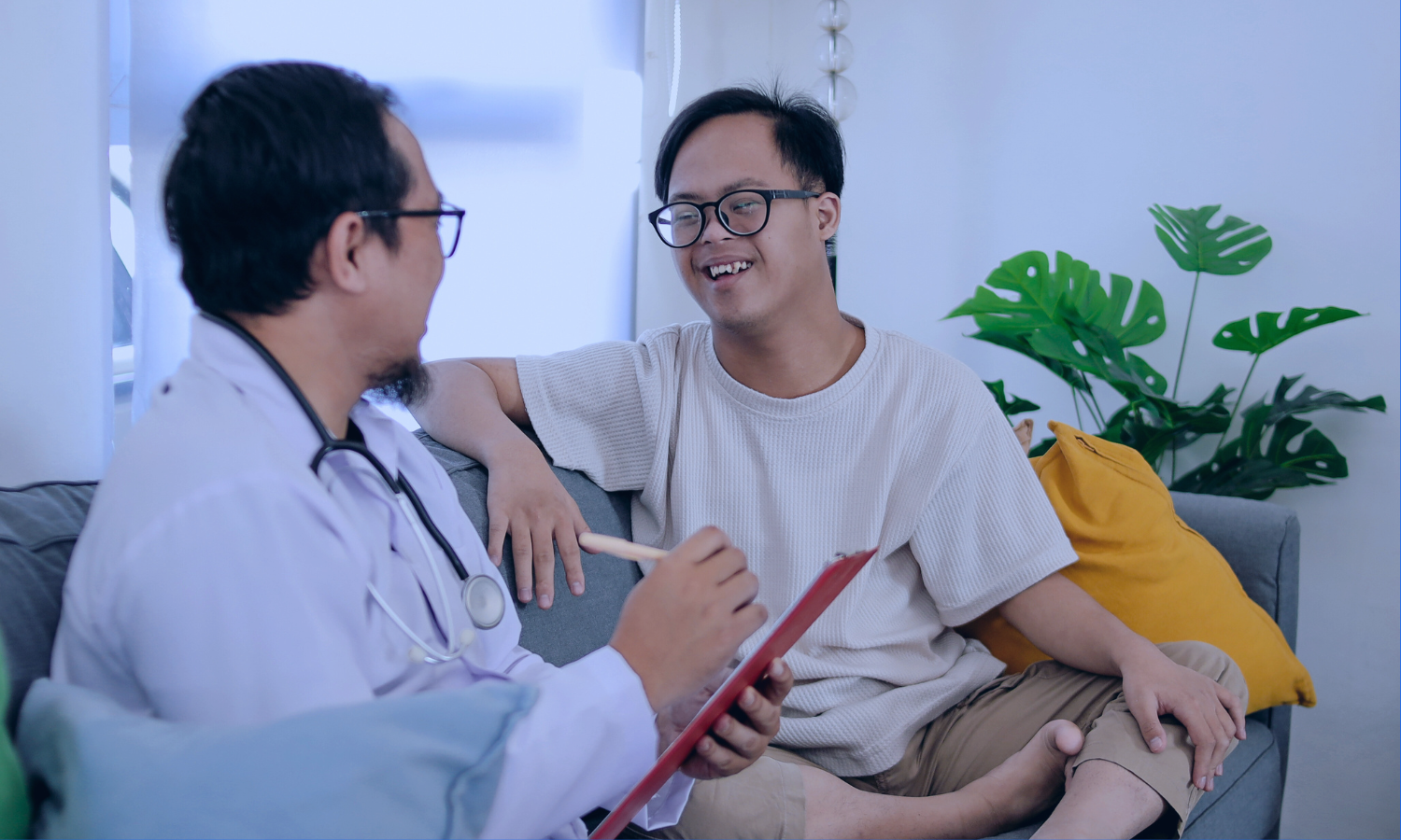 Image of a young boy in a hospital bed hugging his nurse in a comforting embrace. The nurse, wearing a light blue uniform, smiles warmly at the child, expressing care and support. They are in a brightly lit hospital room with large windows in the background, adding a serene and hopeful atmosphere to the scene.